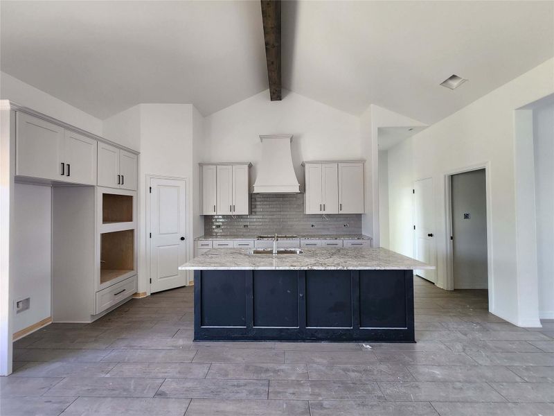 Open-concept kitchen featuring a dark-toned island with a light-toned countertop, white cabinetry with black hardware, a tiled backsplash, and vaulted ceilings with an exposed beam
