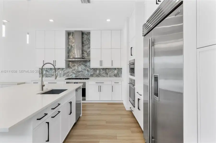 Kitchen featuring white cabinetry, wood-finish flooring, a herringbone-patterned stone backsplash, stainless steel appliances, and a quartz-topped center island