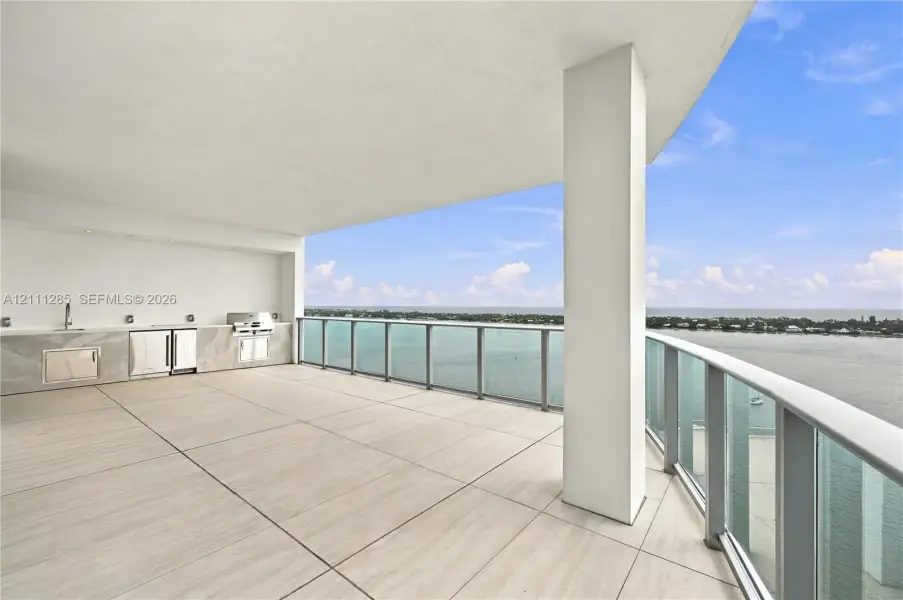 Covered terrace featuring large-format tile flooring, a built-in outdoor kitchen with stone-finish cabinetry, an integrated grill, and a glass-paneled railing Covered terrace featuring large-format tile flooring, a built-in outdoor kitchen with stone-finish cabinetry, an integrated grill, and a glass-paneled railing