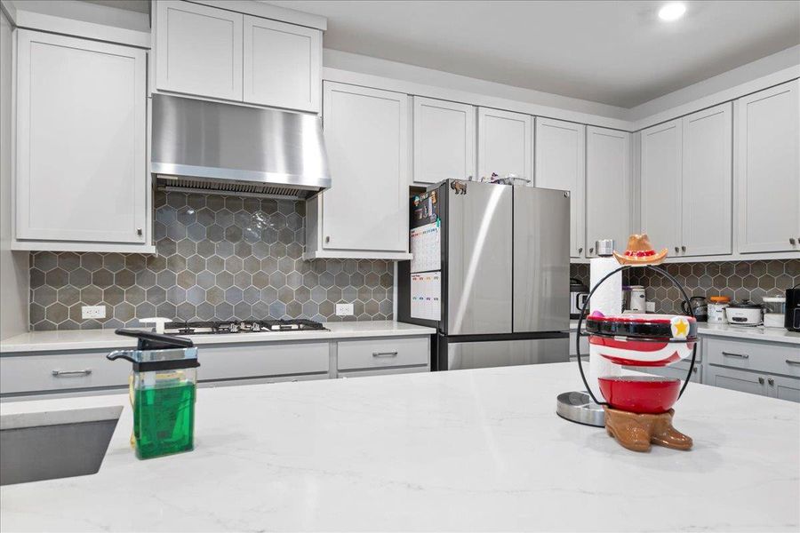 Kitchen featuring freestanding refrigerator, under cabinet range hood, light stone counters, decorative backsplash, and gray cabinets