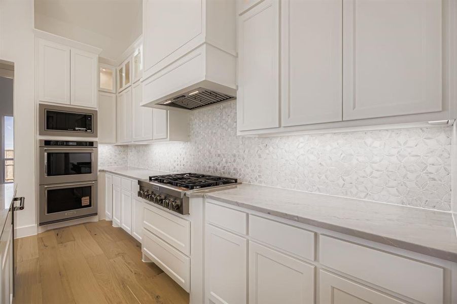 Kitchen with white cabinetry, backsplash, light wood-type flooring, and stainless steel appliances Kitchen with white cabinetry, backsplash, light wood-type flooring, and stainless steel appliances