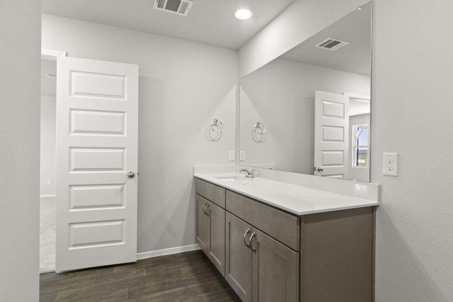 Image of a primary bathroom with brown cabinets, a white vanity, a large mirror and light grey walls