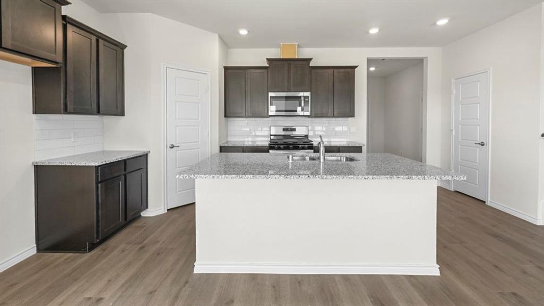 Kitchen with light stone countertops, stainless steel appliances, dark brown cabinetry, backsplash, and a center island with sink