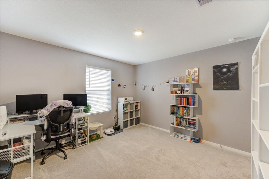 This room features a cozy, carpeted space with neutral walls and ample natural light from a window. It's currently set up as a functional home office with a desk, computer, and shelving for books and decor.