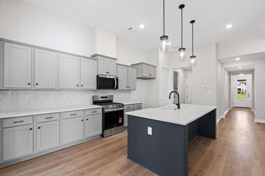 Kitchen featuring light gray cabinetry, stainless steel appliances, a white tile backsplash, a dark island with a built-in sink, and wood-finish flooring Kitchen featuring light gray cabinetry, stainless steel appliances, a white tile backsplash, a dark island with a built-in sink, and wood-finish flooring