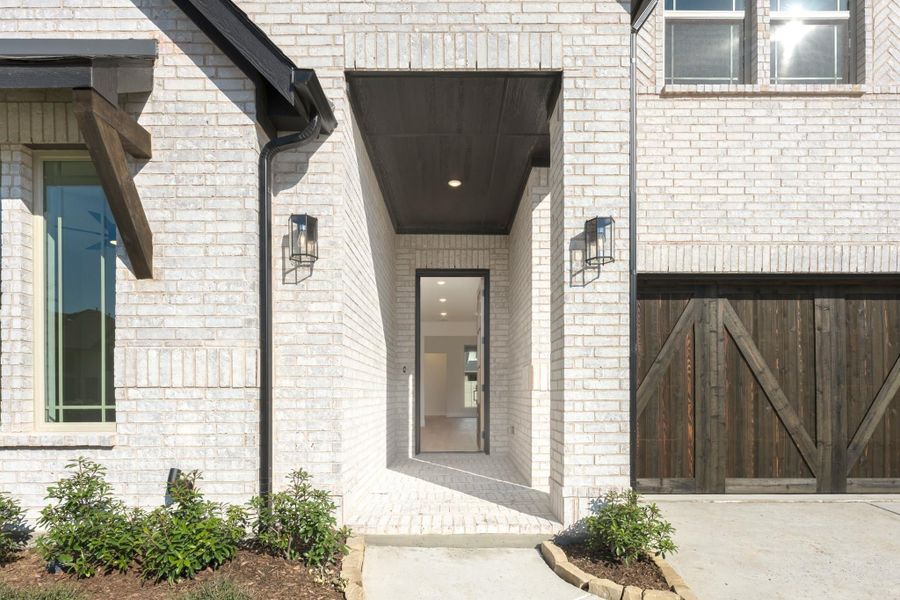 Home entrance with white brick exterior, dark wood covered porch, wall lanterns, and rustic barn-style garage door nearby.