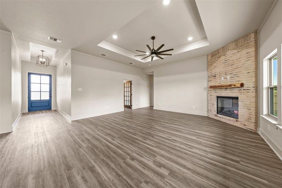 Spacious living area featuring wood-finish flooring, a tray ceiling with recessed lighting and a ceiling fan, a brick-clad fireplace with a wood mantle, and a bright blue front door with glass panels Spacious living area featuring wood-finish flooring, a tray ceiling with recessed lighting and a ceiling fan, a brick-clad fireplace with a wood mantle, and a bright blue front door with glass panels