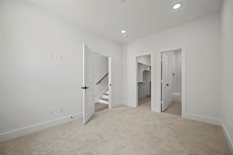 This first level bedroom shows a bright, carpeted room with a neutral color palette. It features three doors: one leading to stairs, another to a spacious walk-in closet, and the third to a modern bathroom with tile flooring. Recessed lighting adds a contemporary touch.