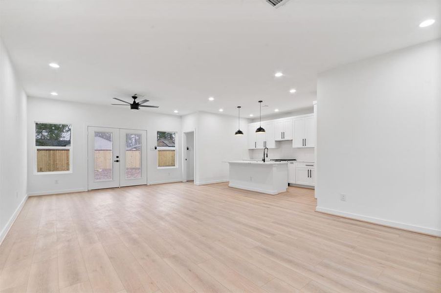 Bright, open-concept living space with light wood flooring and recessed lighting. Features a modern kitchen with white cabinetry, a large island, and black pendant lights. French doors provide backyard access.