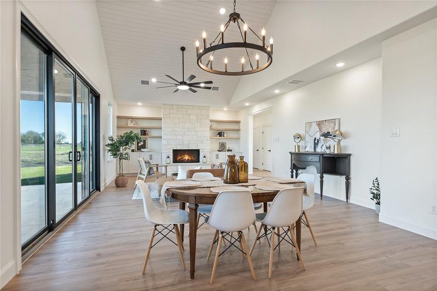 Dining area featuring light wood-type flooring, ceiling fan with notable chandelier, built in features, high vaulted ceiling, and a stone fireplace