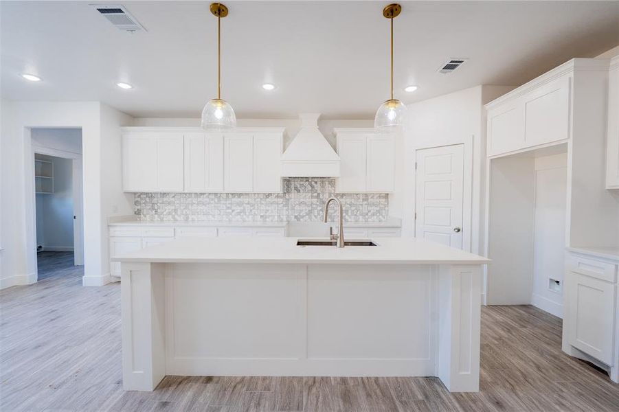 Kitchen with decorative backsplash, pendant lighting, white cabinets, light wood-style flooring, and a center island with sink Kitchen with decorative backsplash, pendant lighting, white cabinets, light wood-style flooring, and a center island with sink