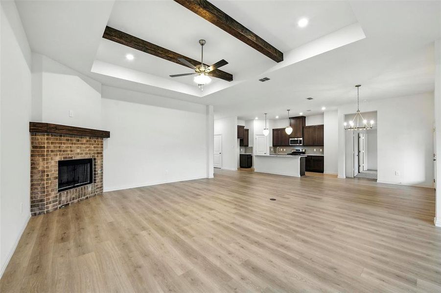 Unfurnished living room featuring ceiling fan, a chandelier, a brick fireplace, light wood finished floors, and beamed ceiling
