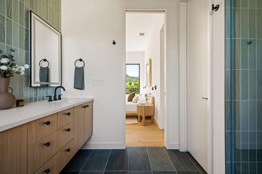 A modern bathroom features a wood vanity, tiled accents, and views into the adjacent bedroom.