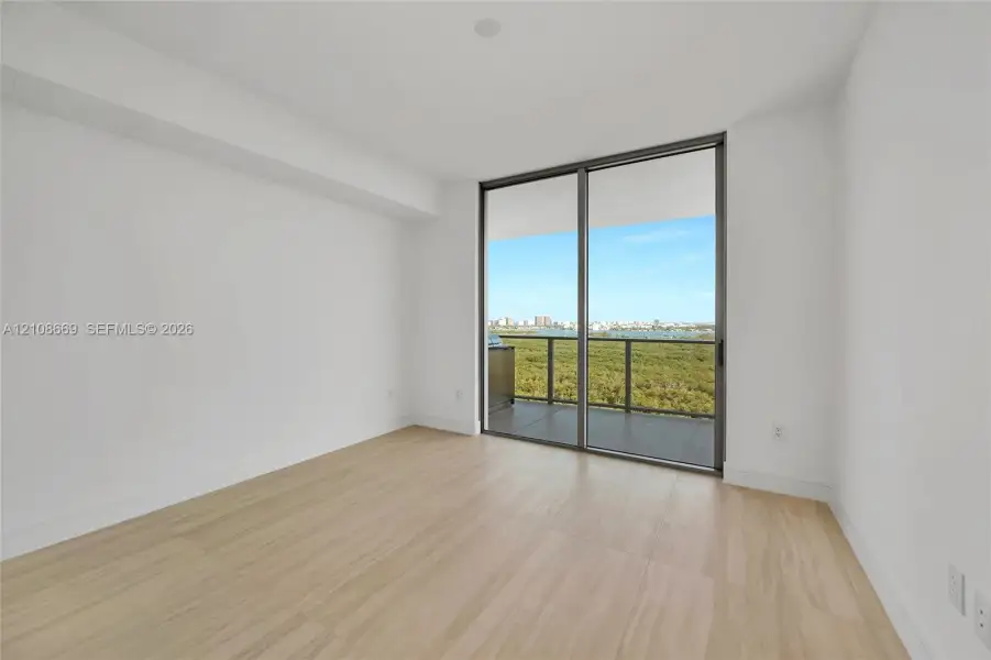 Neutral-toned interior featuring wood-finish flooring, full-height floor-to-ceiling glass sliding doors, and a private balcony with water and coastal scenery