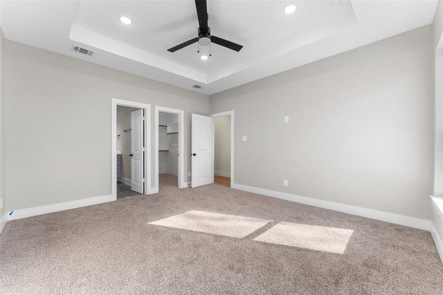 Unfurnished bedroom with ensuite bath, a spacious closet, a tray ceiling, carpet flooring, and ceiling fan