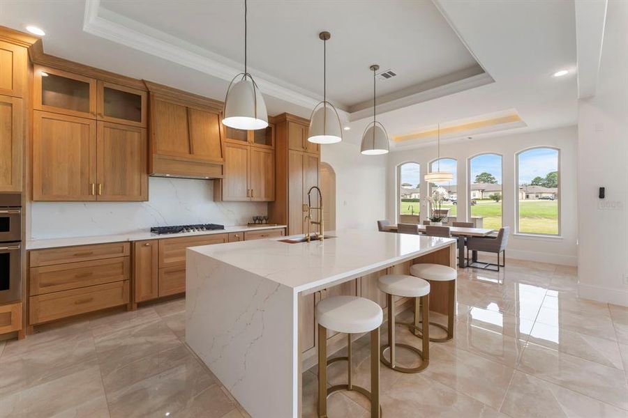 The kitchen opens up to a bright dining area with a view of the outdoors through large arched windows, all under an elegant tray ceiling with contemporary pendant lighting. The bar stools by Star Furniture are absolutely perfect for the space.