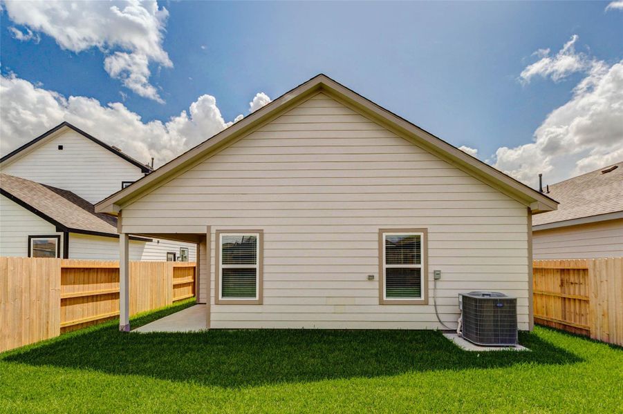 Straight-on rear exterior view highlighting the home’s clean siding, two rear windows, HVAC unit, wood privacy fencing, and grassy backyard. The covered patio is visible to the side, creating a useful outdoor extension of the home while the fully fenced yard provides privacy and everyday convenience.