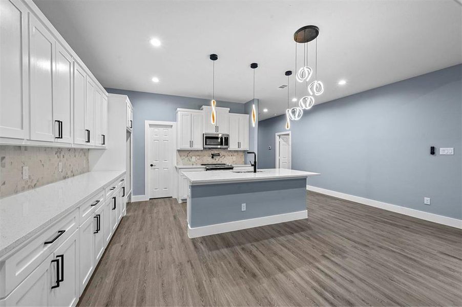 Kitchen featuring hanging light fixtures, light wood-type flooring, stainless steel appliances, white cabinetry, and an island with sink