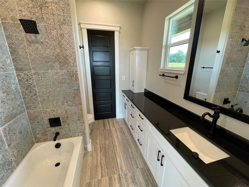 Bathroom featuring a deep soaking tub with a rain shower head, extensive vanity with a black countertop and undermount sink, built-in white cabinetry, wood-finish flooring, and a large mirror