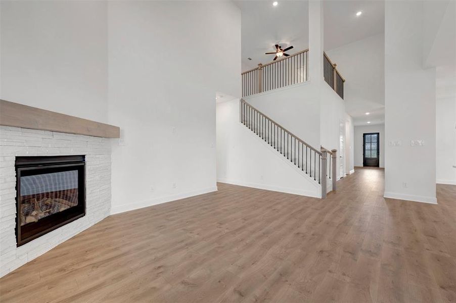 Spacious living area featuring light wood flooring, a fireplace with a white stone surround and wood mantel, and a prominent staircase with dark balusters and light wood handrails