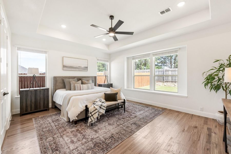 Bedroom with tray ceiling, ceiling fan, hardwood floors, and large windows overlooking a backyard