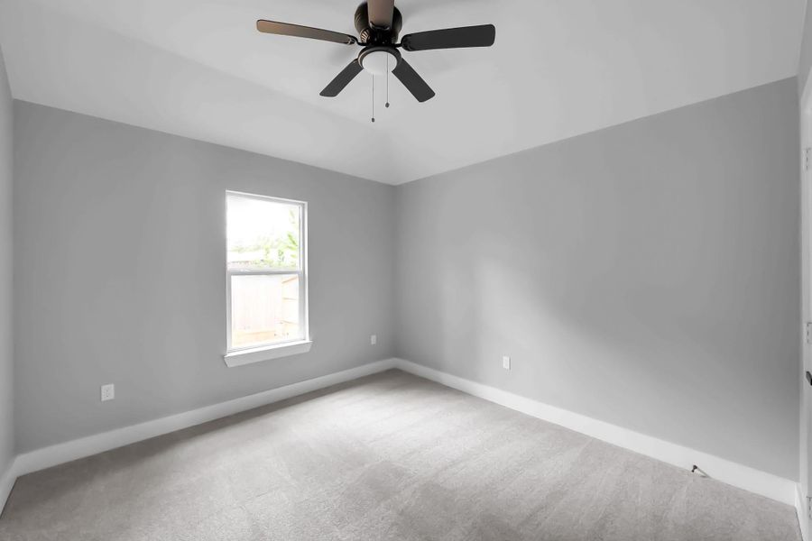 This room features neutral gray walls, a ceiling fan, and a single window allowing natural light, creating a bright and airy space. The carpeted floor adds comfort, making it ideal for a bedroom or office.