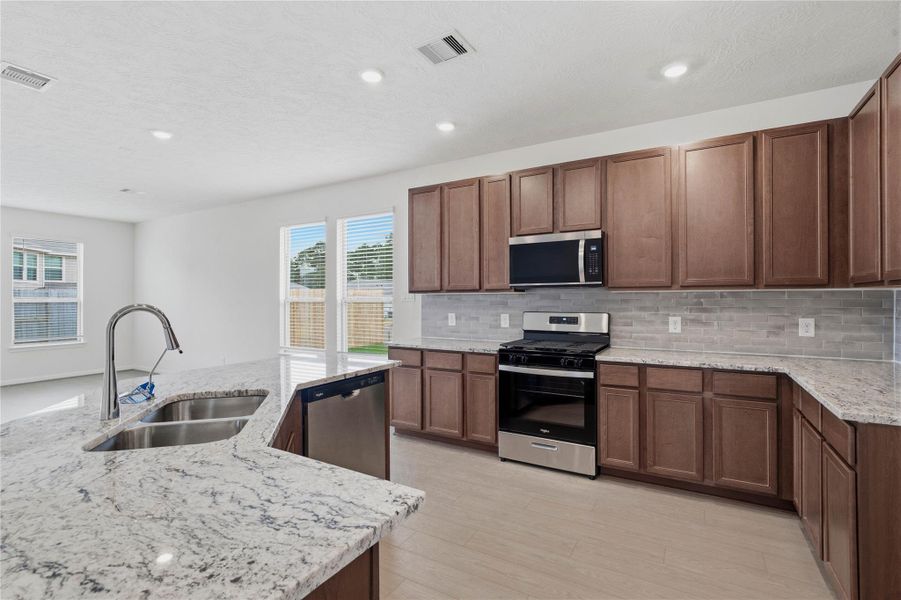 This spacious kitchen boasts high ceilings, Woodmont Cody cabinets in a Ginger stain, Granite White Ice countertops, stainless steel Whirlpool appliances, a modern Daltile backsplash, recessed lighting, windows with blinds, an island, and a pantry—all overlooking the expansive Great Room. This spacious kitchen boasts high ceilings, Woodmont Cody cabinets in a Ginger stain, Granite White Ice countertops, stainless steel Whirlpool appliances, a modern Daltile backsplash, recessed lighting, windows with blinds, an island, and a pantry—all overlooking the expansive Great Room.