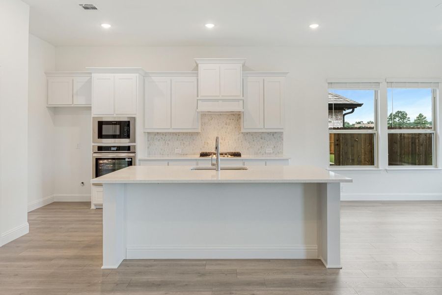 Kitchen with large white island, white cabinets, stone backsplash, and stainless steel appliances on light wood floors