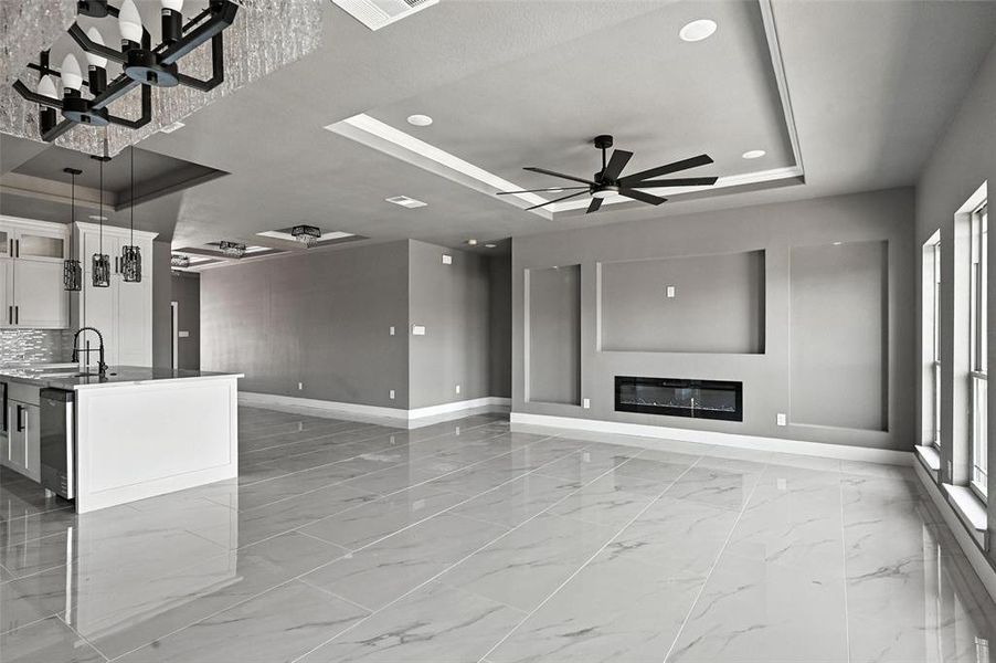 Unfurnished living room featuring a tray ceiling, light marble finish flooring, a glass covered fireplace, a ceiling fan, and healthy amount of natural light Unfurnished living room featuring a tray ceiling, light marble finish flooring, a glass covered fireplace, a ceiling fan, and healthy amount of natural light