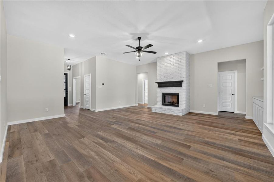 Spacious living area featuring wood-finish flooring, a white brick fireplace with a dark mantel, recessed lighting, a ceiling fan, and built-in cabinetry with open shelving Spacious living area featuring wood-finish flooring, a white brick fireplace with a dark mantel, recessed lighting, a ceiling fan, and built-in cabinetry with open shelving