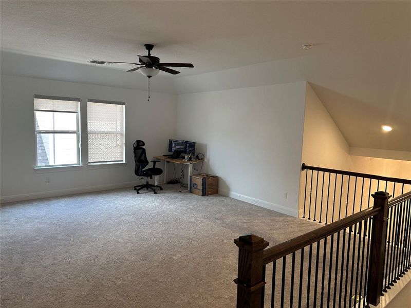 Spacious upper-level room featuring light-colored carpeting, multiple windows with blinds, a ceiling fan with integrated lighting, and a dark wood and metal baluster railing Spacious upper-level room featuring light-colored carpeting, multiple windows with blinds, a ceiling fan with integrated lighting, and a dark wood and metal baluster railing