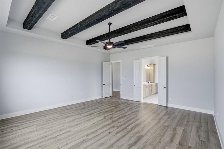 Unfurnished bedroom featuring ceiling fan, ensuite bath, light wood-style floors, and beamed ceiling