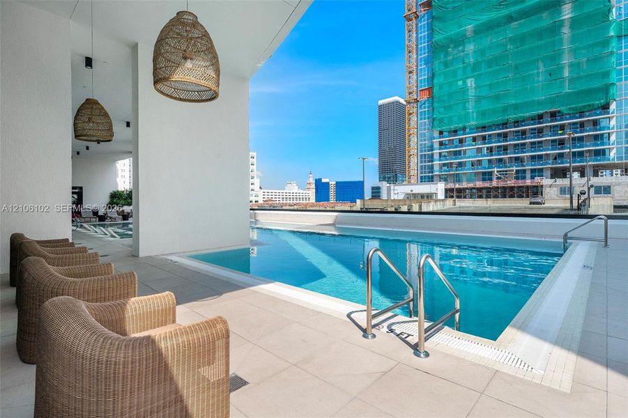 Covered outdoor lounge area featuring large woven pendant lighting, tile flooring, and a rectangular swimming pool with stainless steel railings