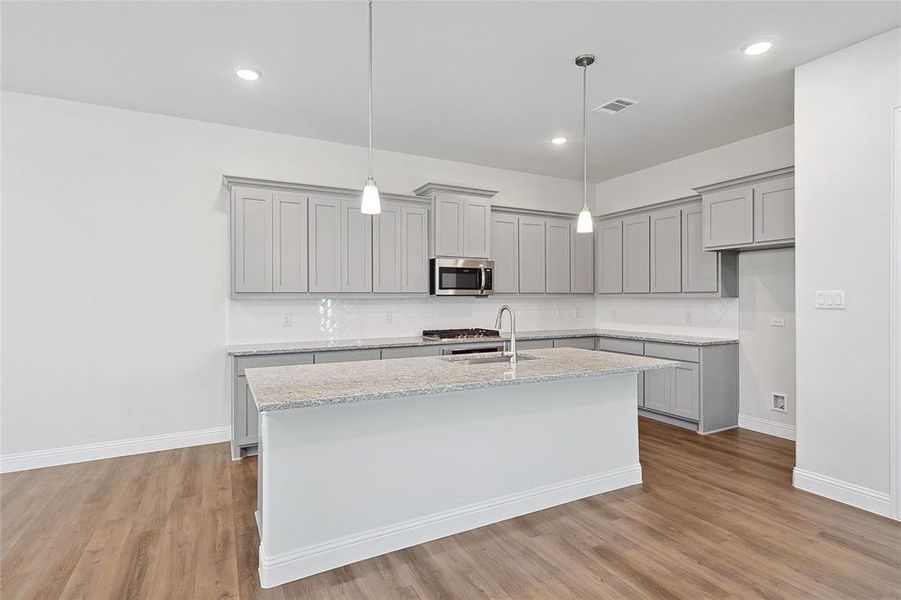 Kitchen with gray cabinets, light stone counters, decorative light fixtures, stainless steel microwave, and an island with sink Kitchen with gray cabinets, light stone counters, decorative light fixtures, stainless steel microwave, and an island with sink