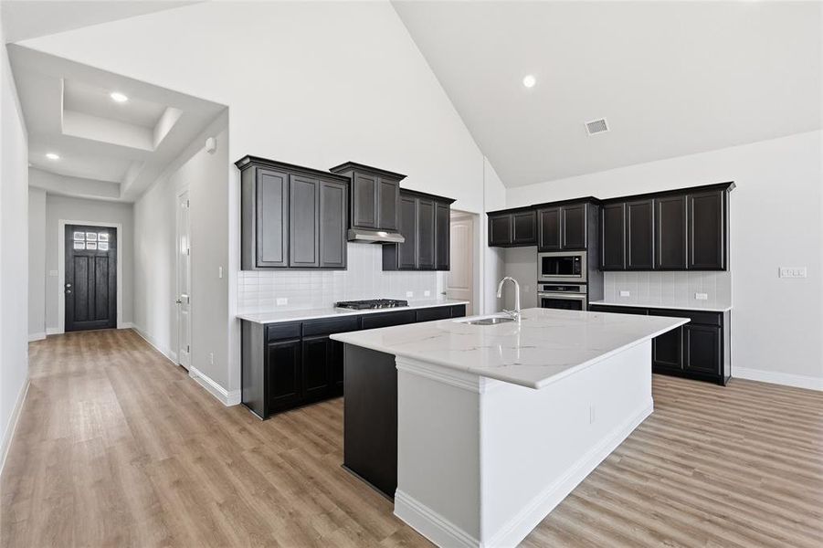 Kitchen with dark cabinets, backsplash, a center island with sink, light stone counters, and vaulted ceiling Kitchen with dark cabinets, backsplash, a center island with sink, light stone counters, and vaulted ceiling