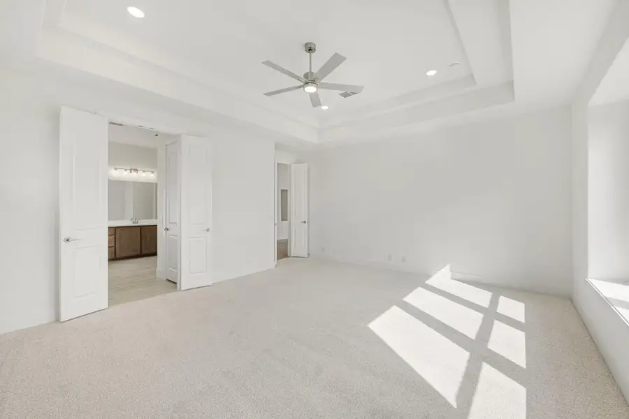 Spacious bedroom with tray ceiling, ceiling fan, carpet flooring, and open door to ensuite bathroom. Spacious bedroom with tray ceiling, ceiling fan, carpet flooring, and open door to ensuite bathroom.