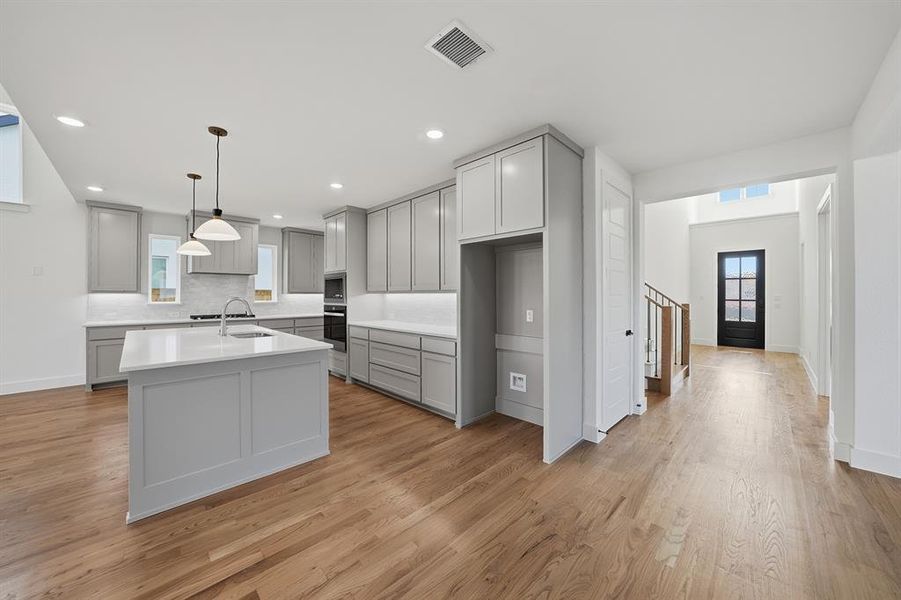 Kitchen featuring gray cabinets, light wood finished floors, pendant lighting, a kitchen island with sink, and decorative backsplash Kitchen featuring gray cabinets, light wood finished floors, pendant lighting, a kitchen island with sink, and decorative backsplash