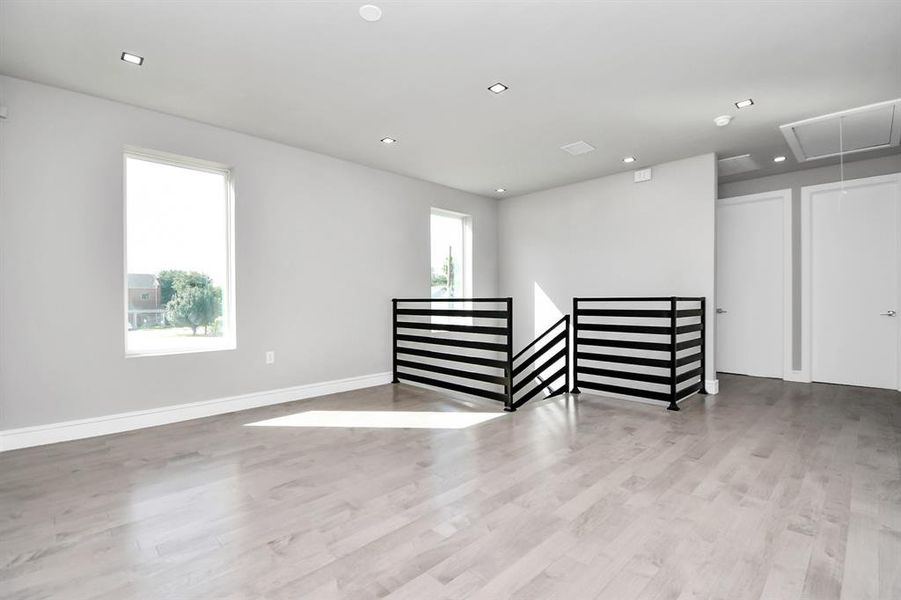 This is a bright, modern room featuring light hardwood floors, fresh gray walls, and contemporary recessed lighting. The space is accented by a stylish black metal railing, and offers multiple doors leading to adjacent rooms, suggesting a versatile layout.