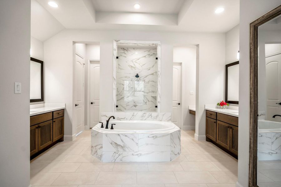This bathroom features a luxurious soaking tub with elegant accents, dual vanities with rich wood cabinetry, and a spacious shower. The neutral color palette and recessed lighting create a bright, inviting atmosphere.