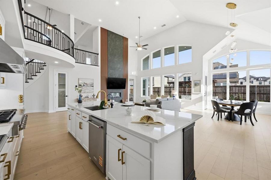 Open-concept living space featuring a gourmet kitchen island with a gold-finish faucet, high vaulted ceilings, an elegant curved staircase with black railings, a contemporary fireplace, and abundant natural light from numerous windows