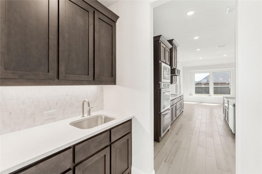 Kitchen with dark wood cabinets, white quartz countertops, stainless sink, tile backsplash, and built-in wall ovens on light wood floors Kitchen with dark wood cabinets, white quartz countertops, stainless sink, tile backsplash, and built-in wall ovens on light wood floors