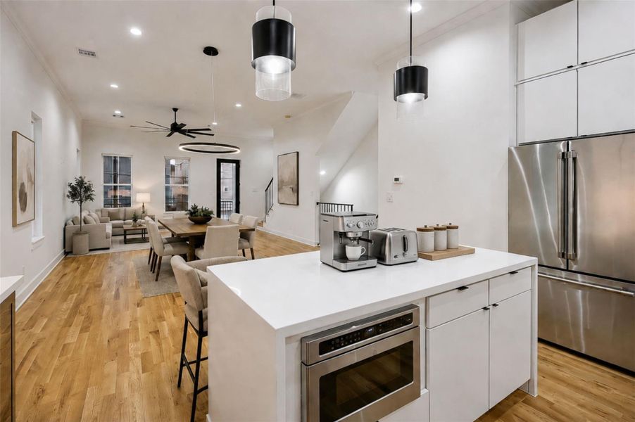 Another kitchen view highlights the spacious island, sleek white cabinetry, stainless steel appliances, and dramatic black pendant lighting. The open layout extends directly into the living and dining areas, creating a highly connected entertaining space. Warm wood flooring adds richness and continuity, while crisp countertops, modern hardware, and abundant cabinetry complete the refined contemporary design. This photo has been virtually staged for visual reference. Another kitchen view highlights the spacious island, sleek white cabinetry, stainless steel appliances, and dramatic black pendant lighting. The open layout extends directly into the living and dining areas, creating a highly connected entertaining space. Warm wood flooring adds richness and continuity, while crisp countertops, modern hardware, and abundant cabinetry complete the refined contemporary design. This photo has been virtually staged for visual reference.