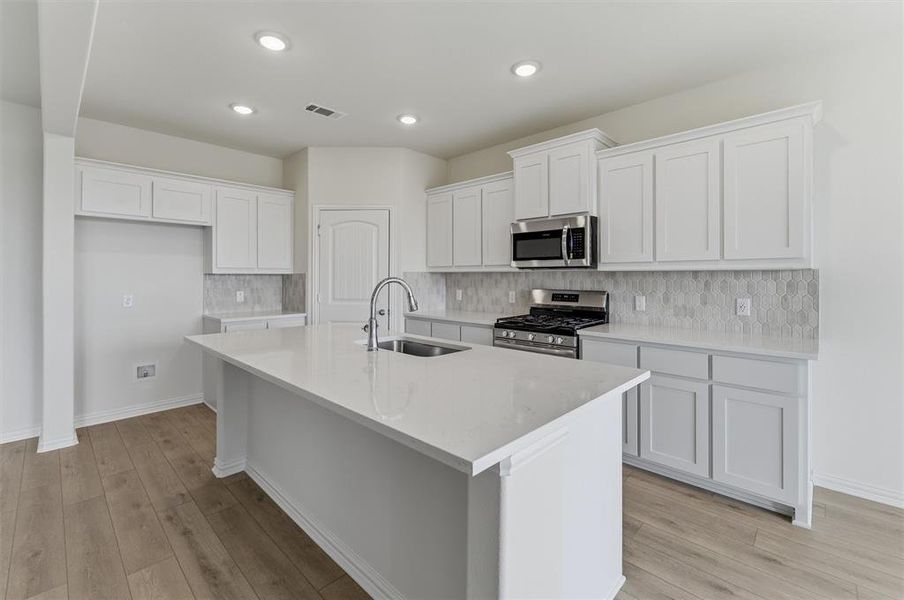 Kitchen featuring a central island with an undermount sink, stainless steel appliances, white cabinetry, a hexagonal tiled backsplash, and wood-finish flooring