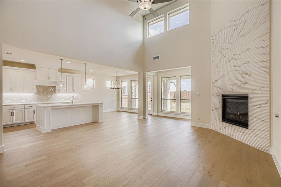 Unfurnished living room with a tile fireplace, light wood-type flooring, a ceiling fan, a towering ceiling, and a chandelier Unfurnished living room with a tile fireplace, light wood-type flooring, a ceiling fan, a towering ceiling, and a chandelier