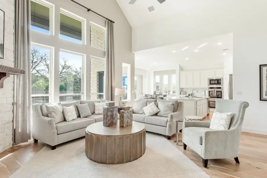 Open-concept living room with vaulted ceiling, tall windows, gray sofas, and kitchen visible in background