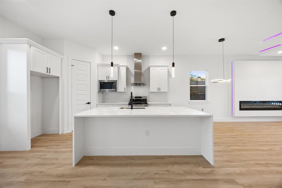 Modern kitchen with a spacious island, sleek white cabinetry, stainless steel appliances, and pendant lighting. The open layout flows into a living area with a contemporary fireplace, creating a bright and inviting space.