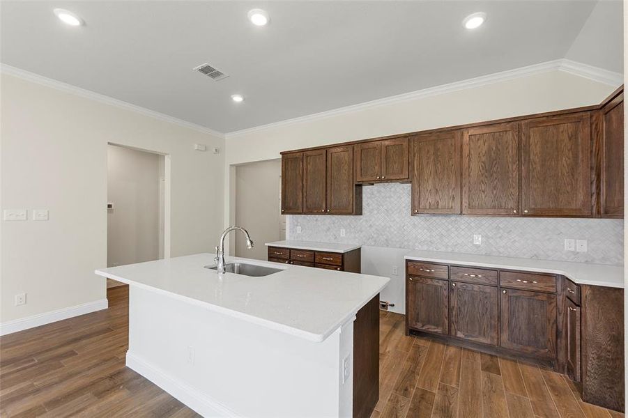 Kitchen with dark wood-type flooring, a center island with sink, sink, ornamental molding, and tasteful backsplash