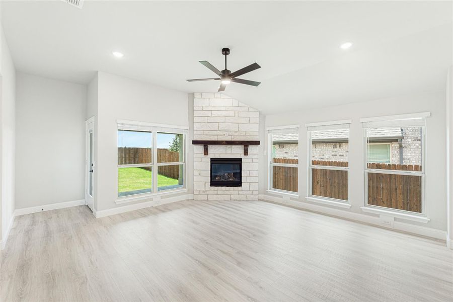 Open living room with stone fireplace, light wood floors, ceiling fan, and large windows