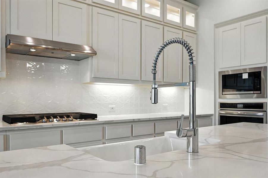 Kitchen with tasteful backsplash, light stone countertops, exhaust hood, appliances with stainless steel finishes, and white cabinetry Kitchen with tasteful backsplash, light stone countertops, exhaust hood, appliances with stainless steel finishes, and white cabinetry