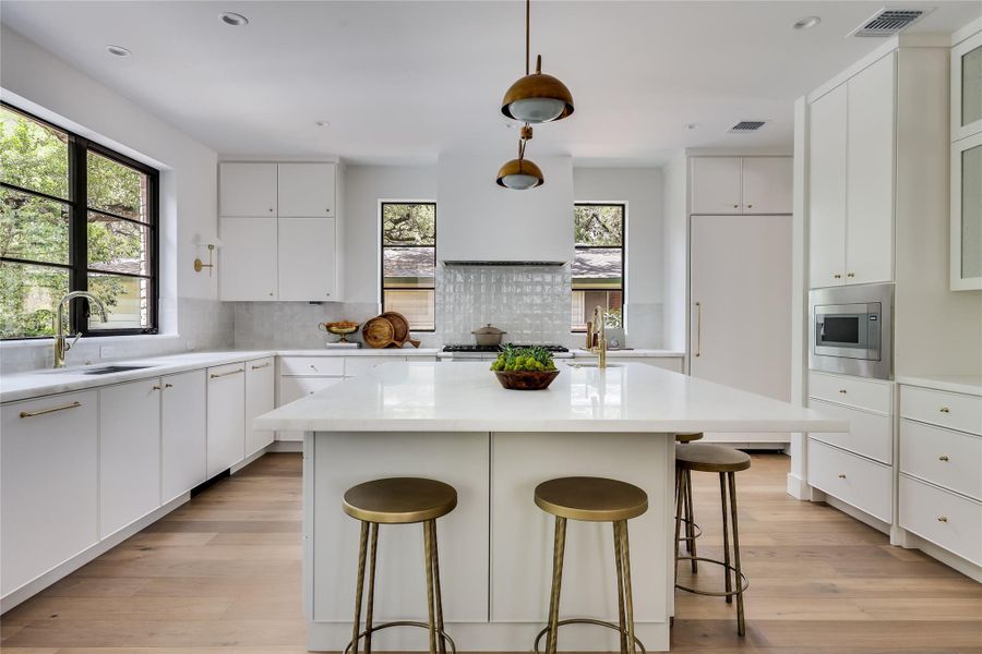 Expansive kitchen featuring a large central island with seating, integrated appliances, wood-finish flooring, white cabinetry with brass hardware, and multiple windows